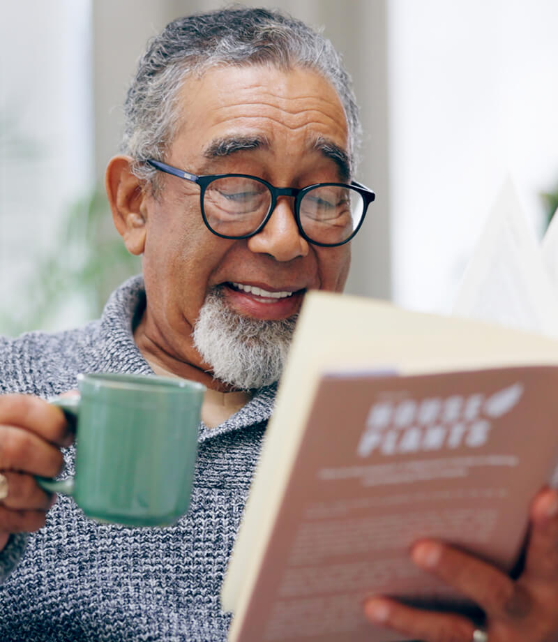 Smiling older man with glasses reading a book and holding a green mug.